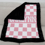 Pink and white checkered blanket with black border and 'Carlynn' text on a gray couch.