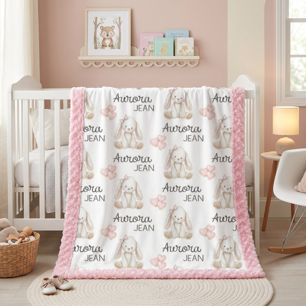 A baby blanket with a white background, pink fuzzy border, and a repeating pattern of gray bunny illustrations and the text 'Aurora JEAN', draped over a white crib in a styled nursery room.