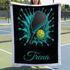 A person holds up a personalized raffle fleece blanket featuring a black background with a teal and black paddle, a yellow-green ping pong ball, and the name 'Trena' in teal script. The blanket has a white plush border and is photographed on a tennis court.