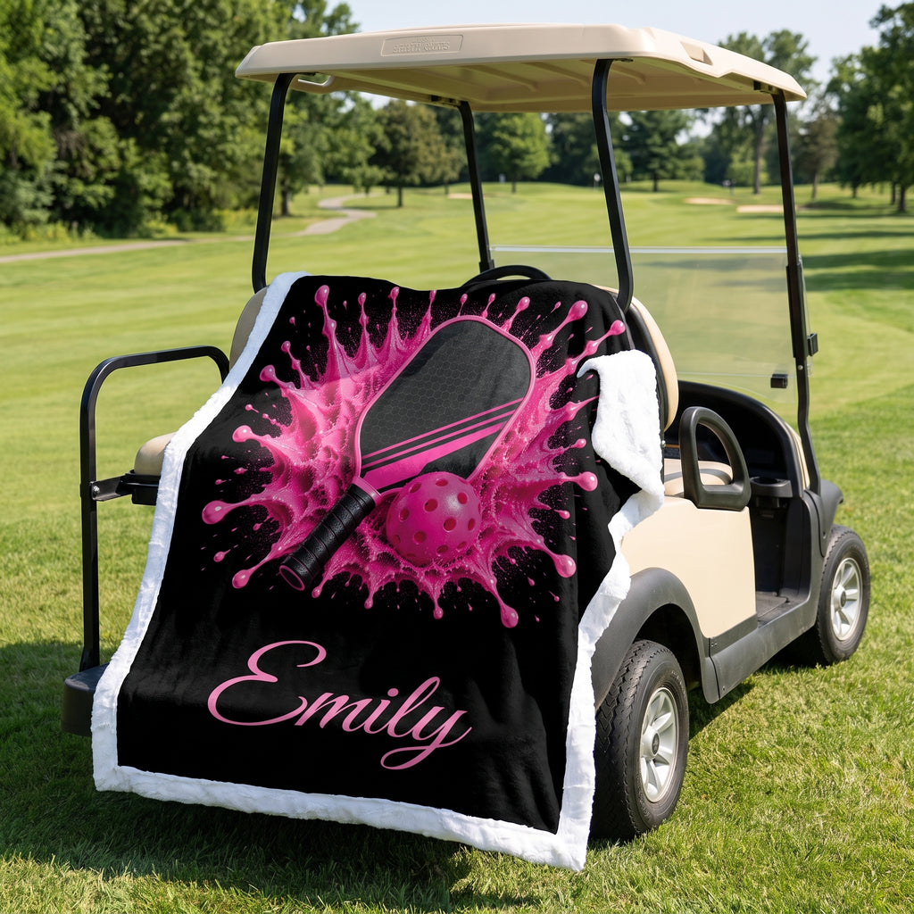 A custom golf-themed throw blanket draped over a beige golf cart on a grassy golf course. The blanket features a black background with a vibrant pink pickleball splash graphic, a pink paddle, a pink ball, and the name 'Emily' in pink script at the bottom.