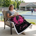 A woman sits in a patio chair on a tennis court, reading a book while wrapped in a black fleece blanket with a pink paint splatter design, a paddle, a ball, and the name 'Emily' in pink script.