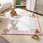 A personalized baby milestone play mat with a pink gingham border, featuring a knitted elephant illustration, the name 'Eliana Elizabeth,' and a numbered age tracker. The mat is styled in a bright nursery setting with a teddy bear, baby booties, wooden blocks, and toys.