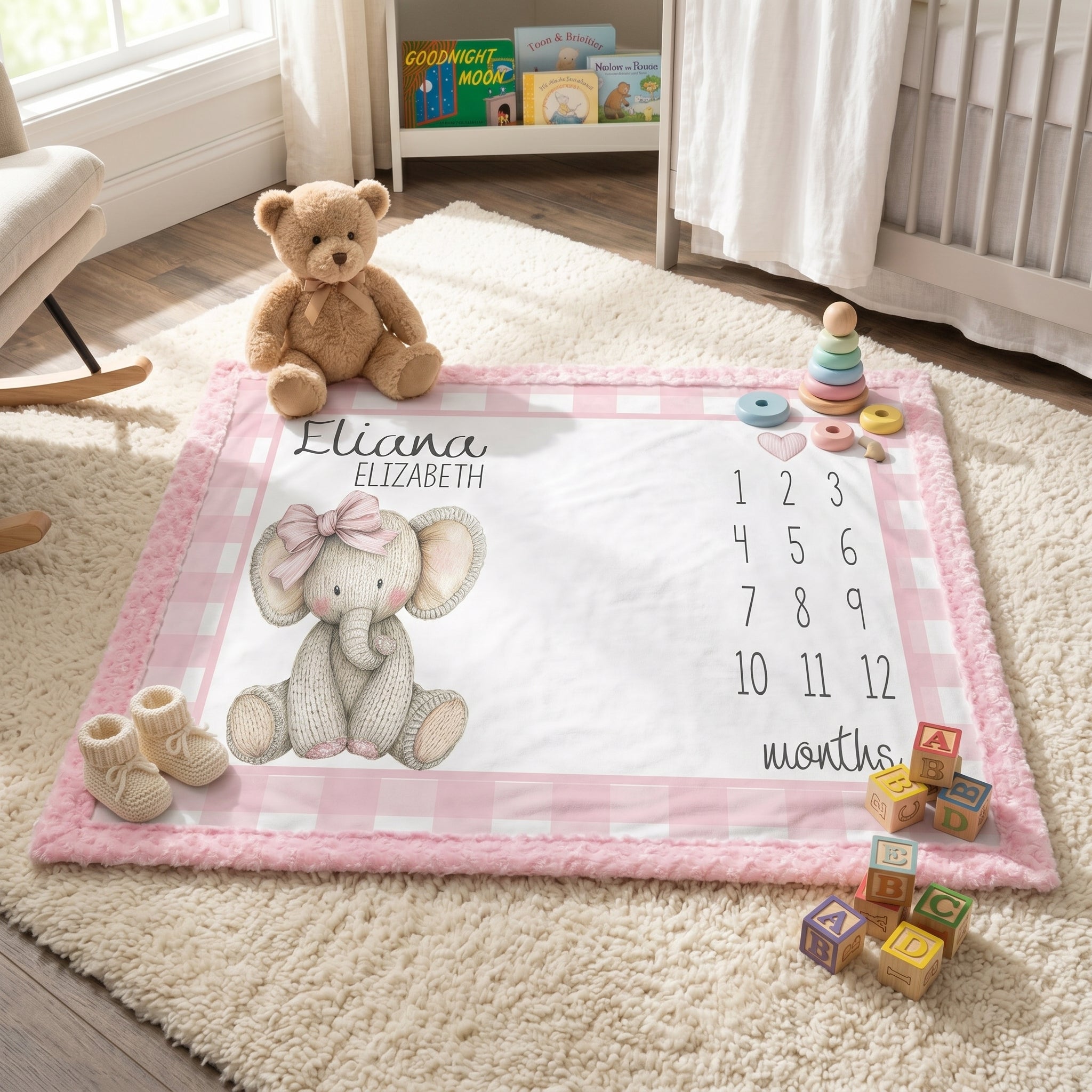 A personalized baby milestone play mat with a pink gingham border, featuring a knitted elephant illustration, the name 'Eliana Elizabeth,' and a numbered age tracker. The mat is styled in a bright nursery setting with a teddy bear, baby booties, wooden blocks, and toys.