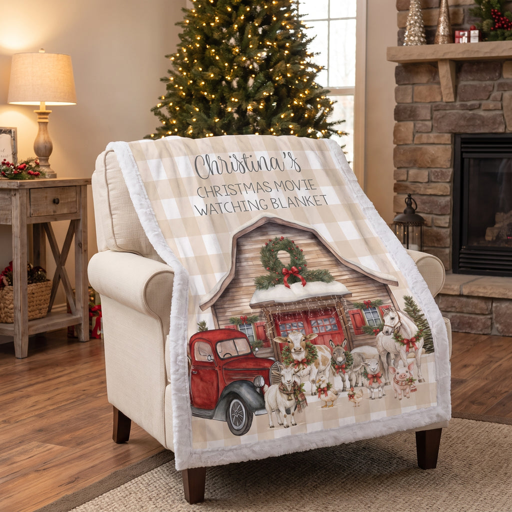 A Christmas-themed fleece throw blanket draped over a beige armchair in a cozy living room. The blanket features a beige and white gingham pattern border, a central illustration of a rustic cabin with a red vintage truck, Santa Claus, and dogs, and the text 'Christmas Movie Watching Blanket' printed at the top.