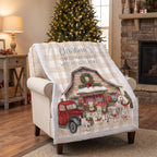 A Christmas-themed fleece throw blanket draped over a beige armchair in a cozy living room. The blanket features a beige and white gingham pattern border, a central illustration of a rustic cabin with a red vintage truck, Santa Claus, and dogs, and the text 'Christmas Movie Watching Blanket' printed at the top.