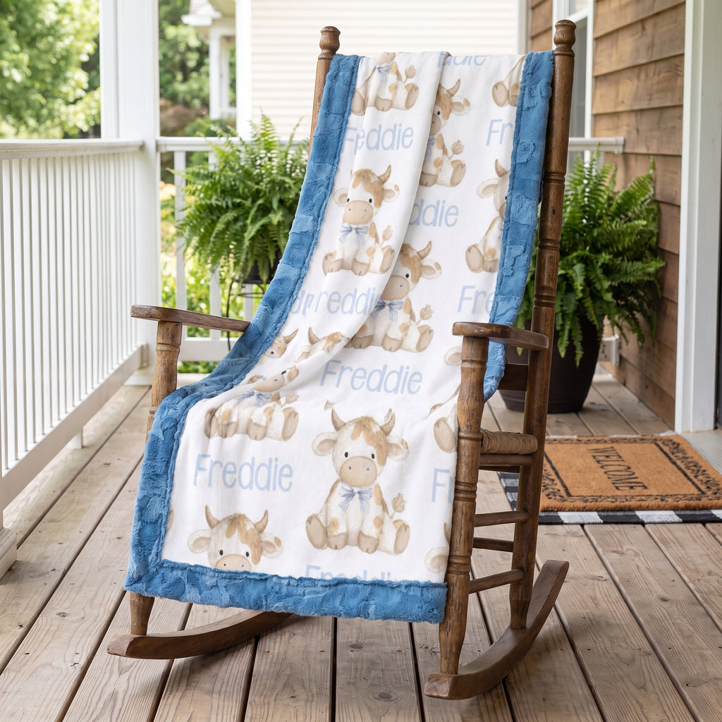 A personalized baby blanket with a blue minky fabric border and a white center printed with teddy bear and cow illustrations, including the name 'Freddie', draped over a wooden rocking chair on a porch.