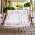 A personalized baby blanket draped over a wooden porch swing, featuring a white center with pink floral borders, bows, and a printed monthly calendar grid labeled 'Clara months'.