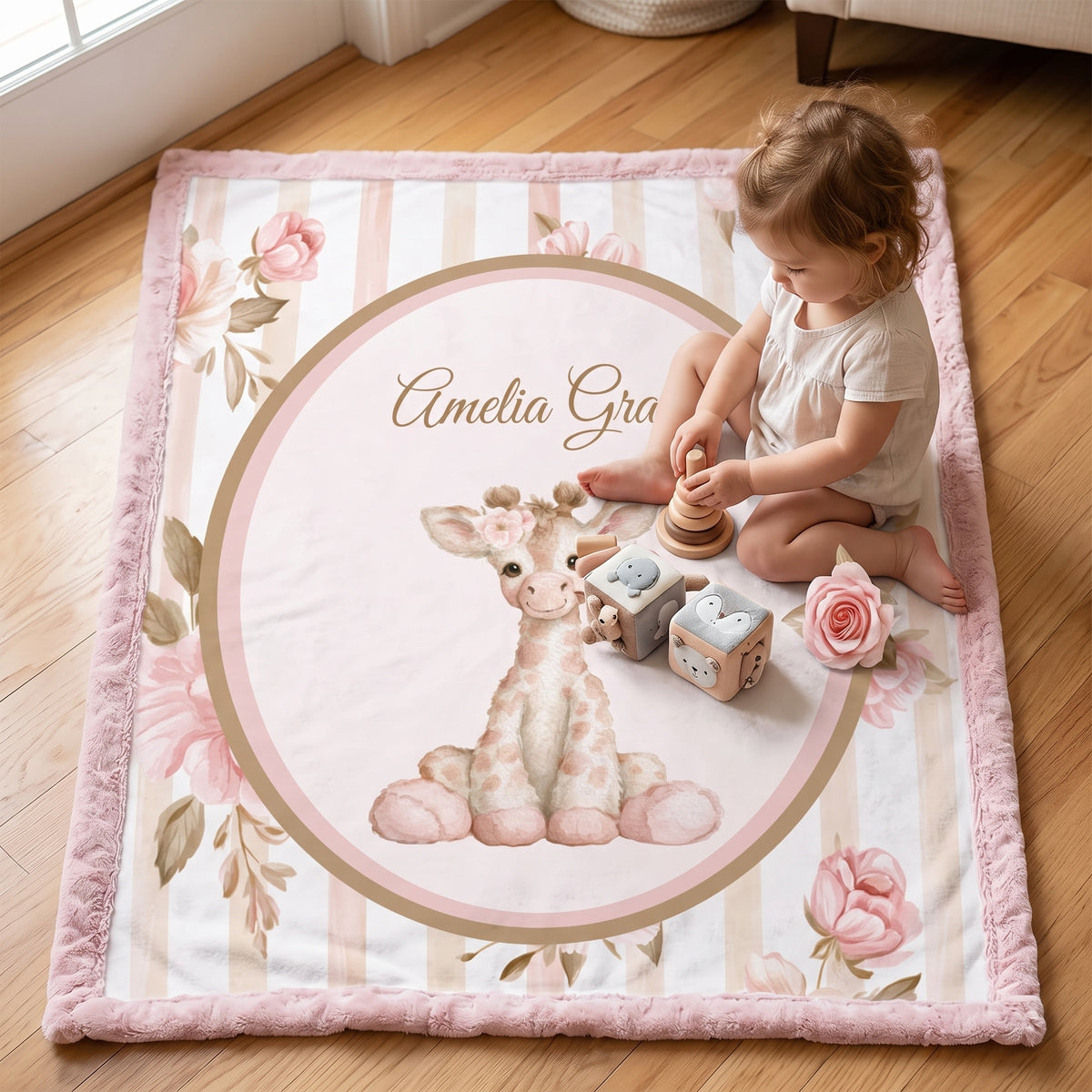 A baby girl's personalized play mat on a wooden floor, featuring a pink giraffe, pink roses, and the name 'Amelia Grace' in a central oval. A toddler is sitting on the mat playing with wooden toys.
