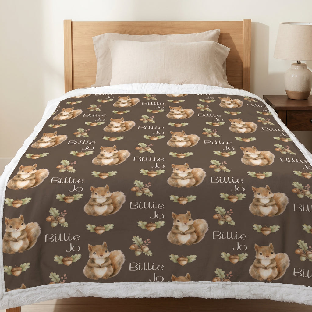 A bedding set on a bed, featuring a dark brown fleece blanket with a repeating pattern of brown and white squirrels, acorns, leaves, and the text 'Billie Jo', paired with a white sherpa fleece throw blanket.