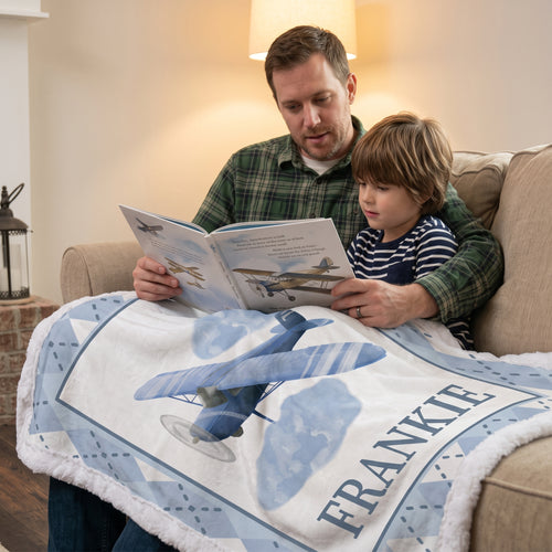 A man and a young boy sit on a beige couch reading a book together, wrapped in a personalized blanket featuring a blue biplane illustration and the name 'FRANKIE' in large letters.