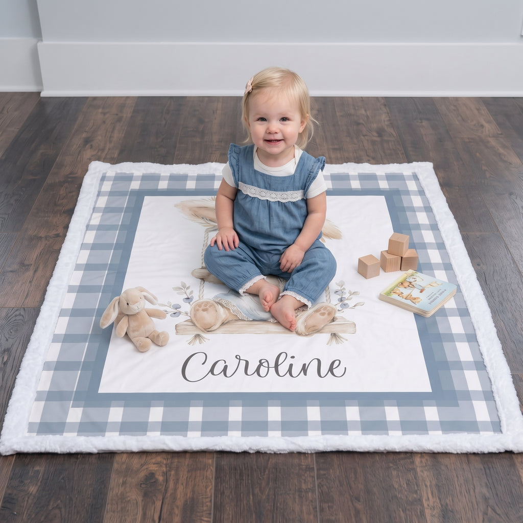 A toddler sits cross-legged on a personalized baby blanket on a dark wood floor. The blanket features a blue and white gingham border, a white center with floral embroidery, and the name 'Caroline' printed at the bottom.