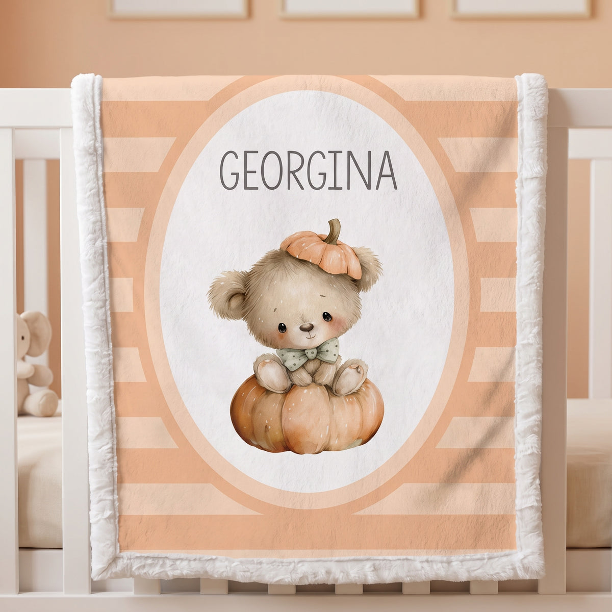 Personalized baby blanket with a cute teddy bear and pumpkin design, featuring the name 'GEORGINA' in the center, draped over a white crib.