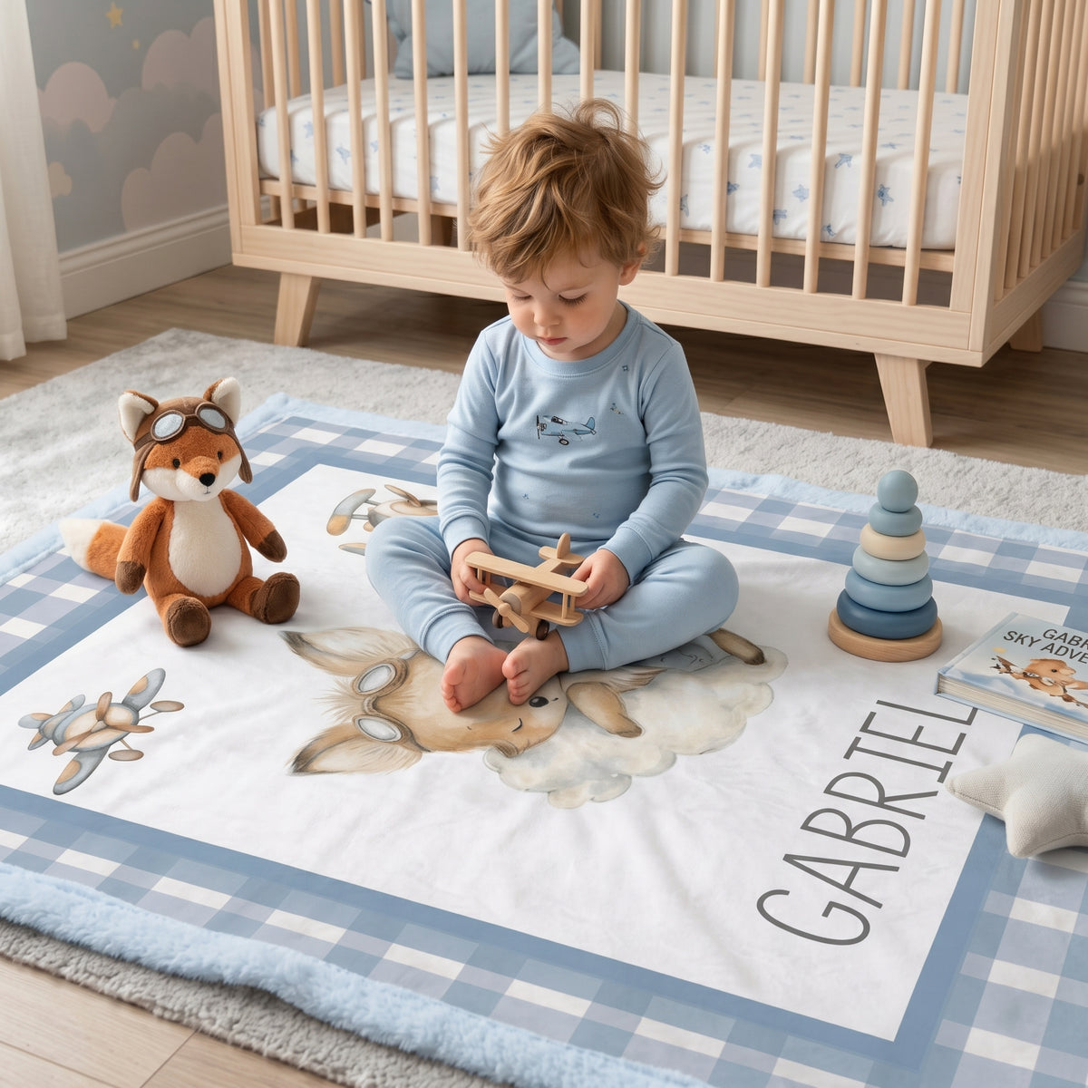 A toddler sits cross-legged on a personalized baby play mat in a nursery, wearing a light blue long-sleeve pajama set. The play mat features a blue and white gingham border, a watercolor deer design, the name 'GABRIEL' printed in black, and is styled with toys including a plush fox and a wooden stacking toy.