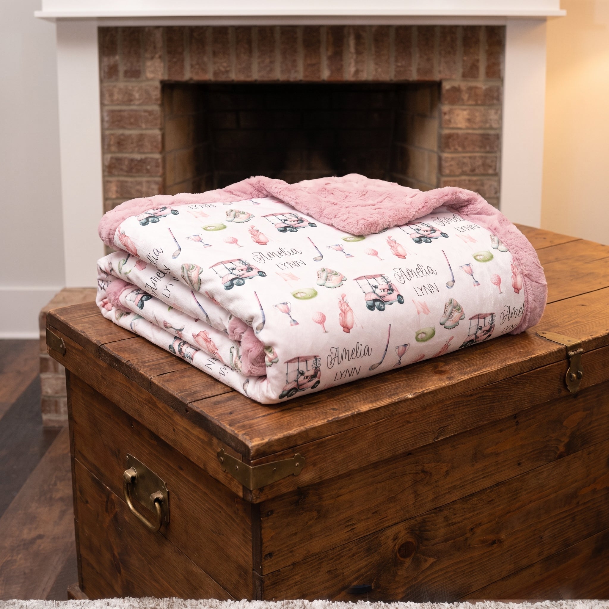 A folded baby blanket with a pink sherpa lining and a white printed pattern featuring baby bottles, stovetops, and the name 'Amanda 1999', displayed on a rustic wooden chest in front of a brick fireplace.