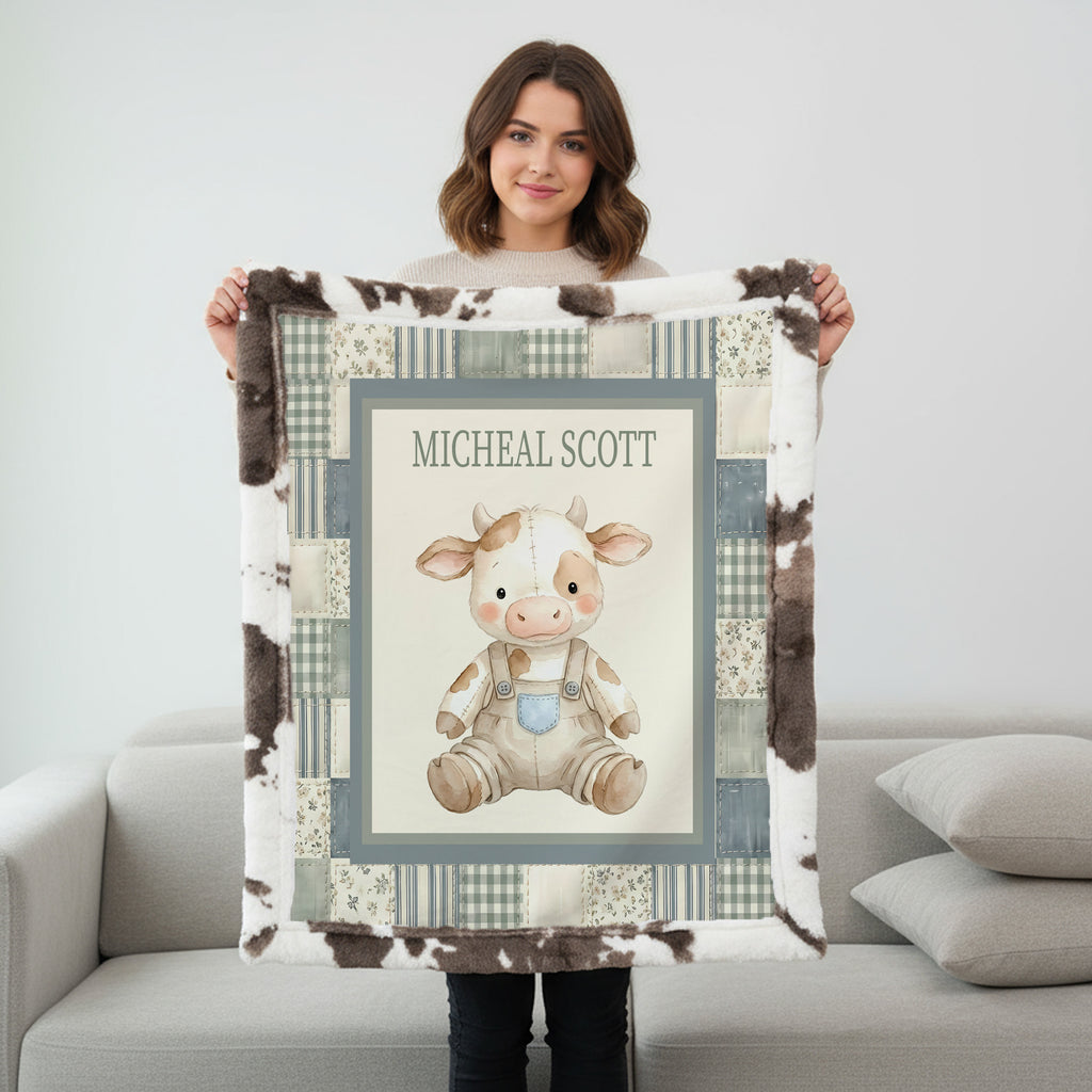 A woman stands in a living room holding up a personalized plush fleece blanket. The blanket features a cow-print border, a central cream panel with a cartoon baby cow illustration, and the name 'MICHEAL SCOTT' printed at the top.