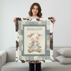 A woman stands in a living room holding up a personalized plush fleece blanket. The blanket features a cow-print border, a central cream panel with a cartoon baby cow illustration, and the name 'MICHEAL SCOTT' printed at the top.