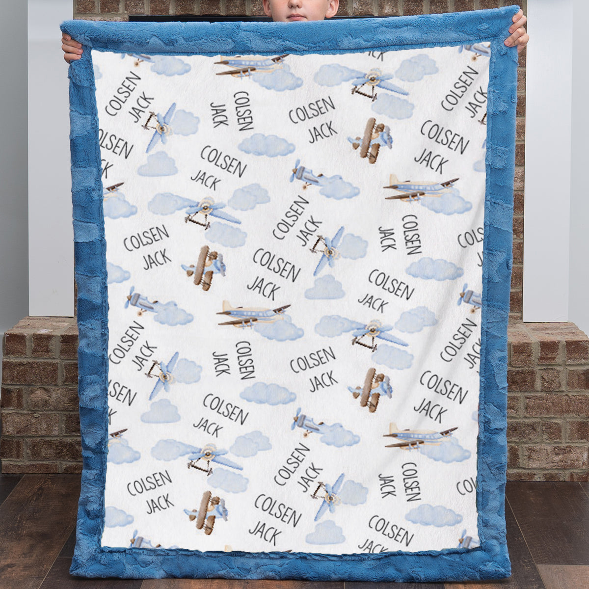 A child holds up a minky blanket featuring a blue border and a white center printed with blue clouds, brown airplanes, and the text 'COLSEN JACK' and 'DON'T Worry'.
