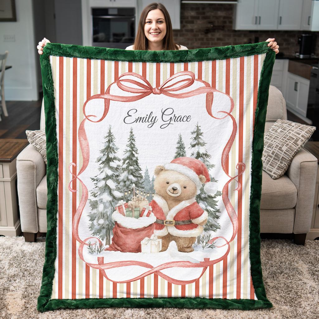 A woman holds up a personalized Christmas-themed fleece blanket in a living room setting. The blanket features a festive design with a green plush border, red and cream vertical stripes, a large red bow, evergreen trees, a teddy bear in a Santa suit, and the custom name 'Emily Grace' printed at the top.