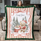 A woman holds up a personalized Christmas-themed fleece blanket in a living room setting. The blanket features a festive design with a green plush border, red and cream vertical stripes, a large red bow, evergreen trees, a teddy bear in a Santa suit, and the custom name 'Emily Grace' printed at the top.