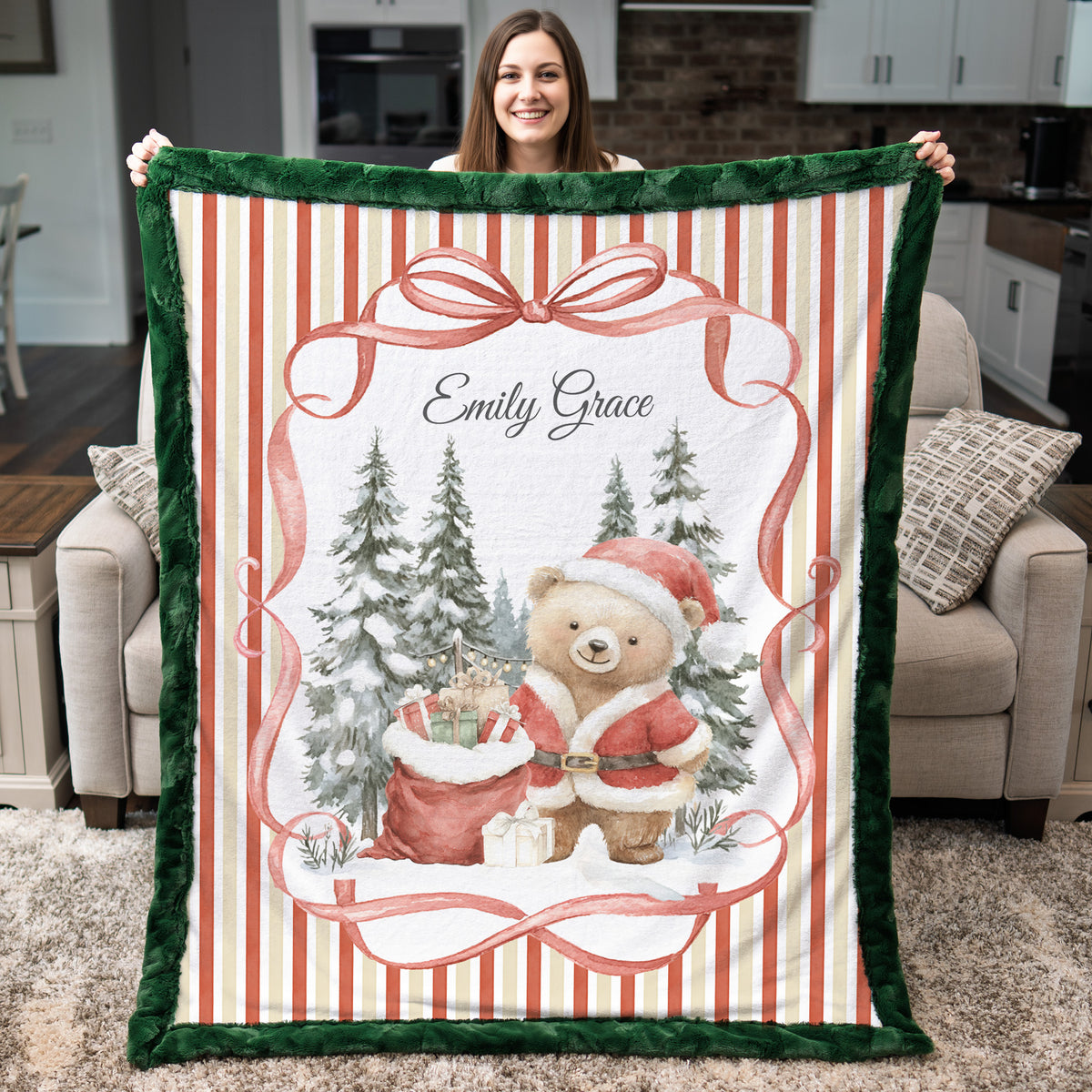 A woman holds up a personalized Christmas-themed fleece blanket in a living room setting. The blanket features a festive design with a green plush border, red and cream vertical stripes, a large red bow, evergreen trees, a teddy bear in a Santa suit, and the custom name 'Emily Grace' printed at the top.