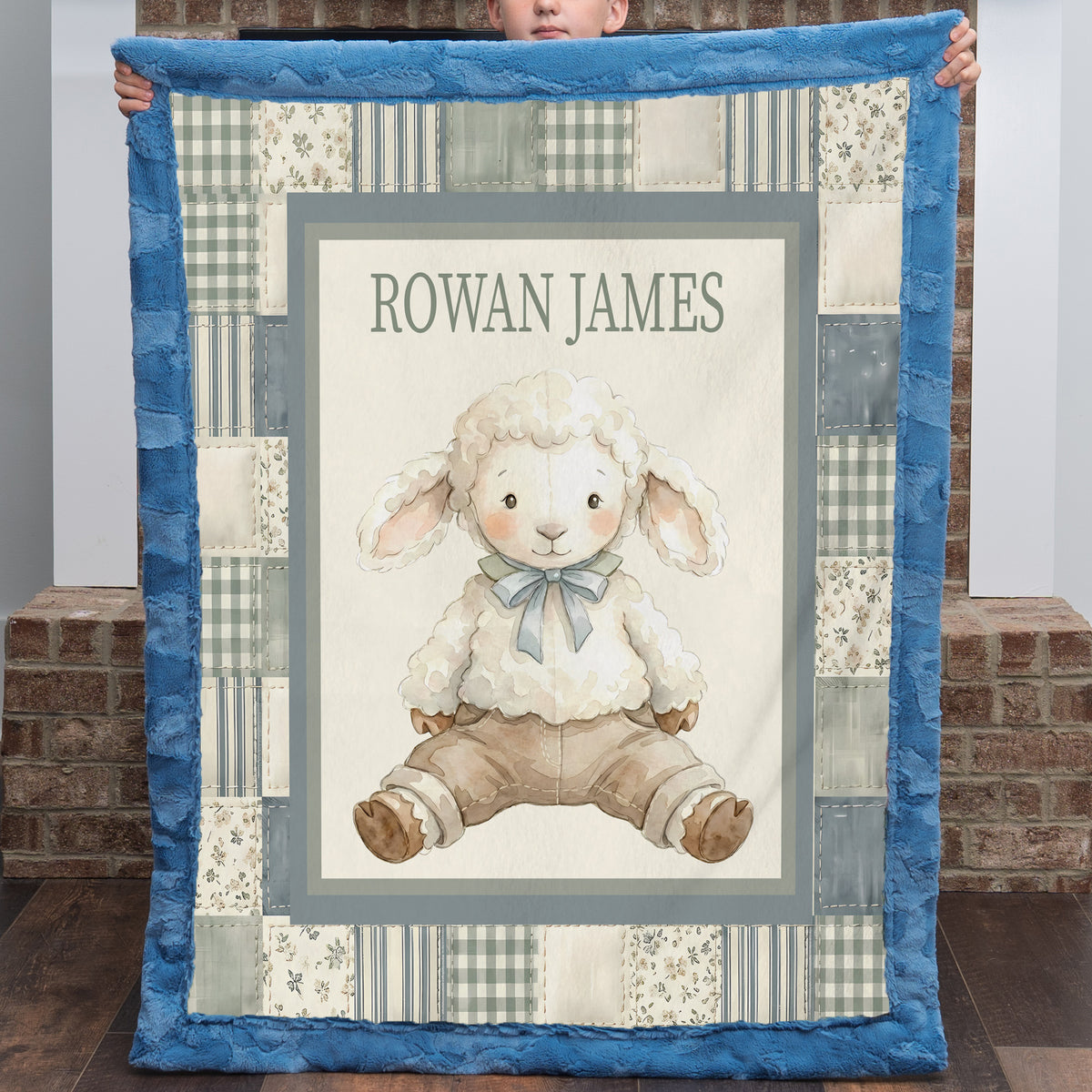 A personalized quilted baby blanket held up by a child, featuring a central illustration of a fluffy white lamb wearing a blue bow and brown pants, surrounded by a blue border and a patchwork design with the name 'ROWAN JAMES' at the top.