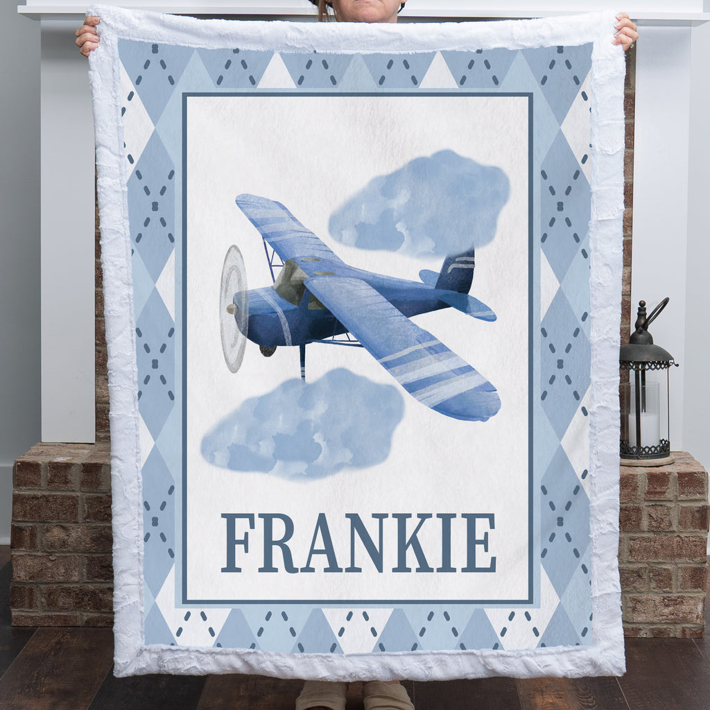 A person holds up a quilted throw blanket featuring a vintage-style blue airplane illustration, two clouds, the name 'FRANKIE' in large letters, and a light blue and white argyle border. The photo is taken indoors in front of a brick column and lantern.