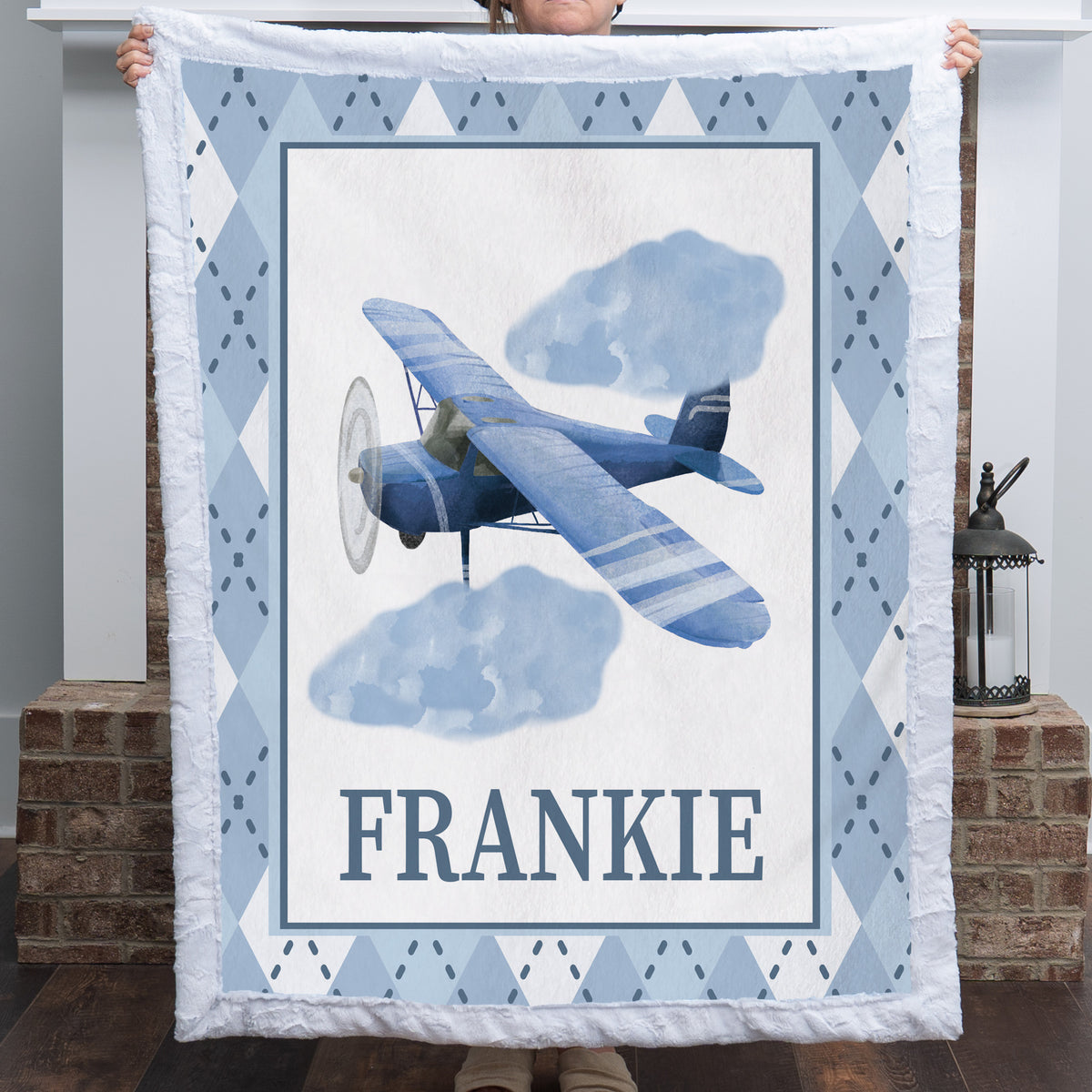 A person holds up a quilted throw blanket featuring a vintage-style blue airplane illustration, two clouds, the name 'FRANKIE' in large letters, and a light blue and white argyle border. The photo is taken indoors in front of a brick column and lantern.