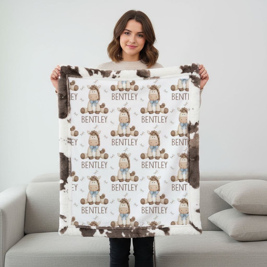 A woman stands in a living room holding up a custom fleece blanket. The blanket features a white background with a repeating pattern of cartoon baby characters wearing blue outfits and brown hats, with the name 'BENTLEY' printed below each character.
