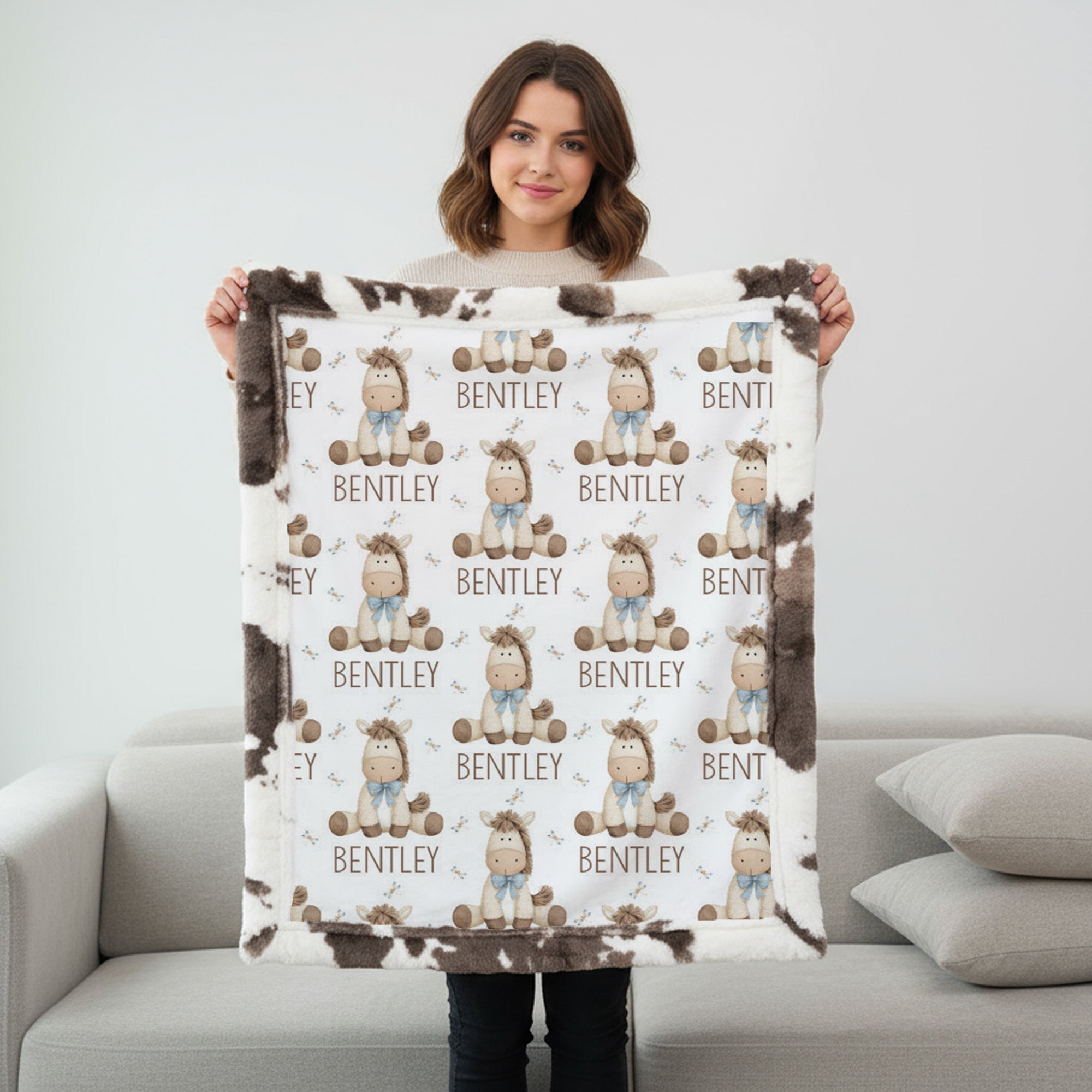 A woman stands in a living room holding up a custom fleece blanket. The blanket features a white background with a repeating pattern of cartoon baby characters wearing blue outfits and brown hats, with the name 'BENTLEY' printed below each character.