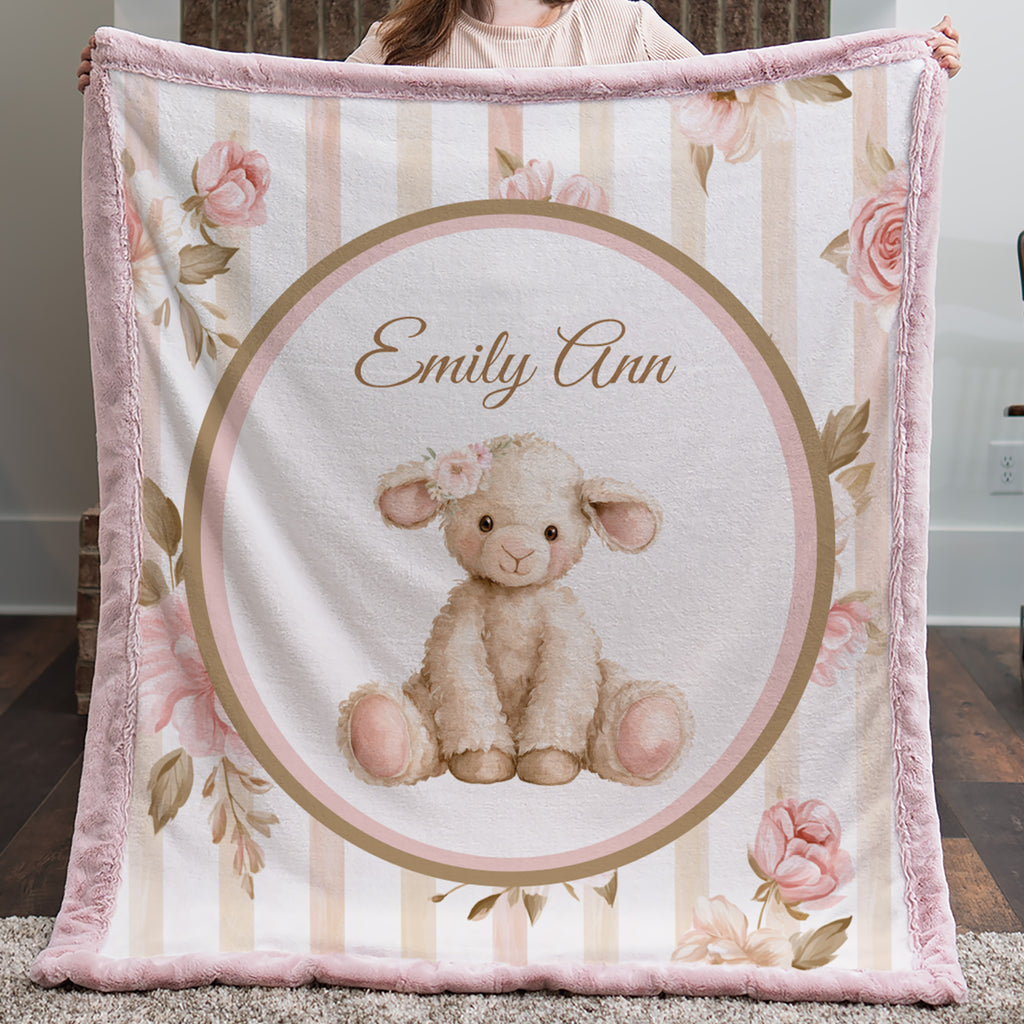 Personalized baby blanket held up by a person, featuring a central illustration of a fluffy cream-colored lamb inside a circular frame with the text 'Emily Ann', surrounded by pink and beige vertical stripes and floral designs.