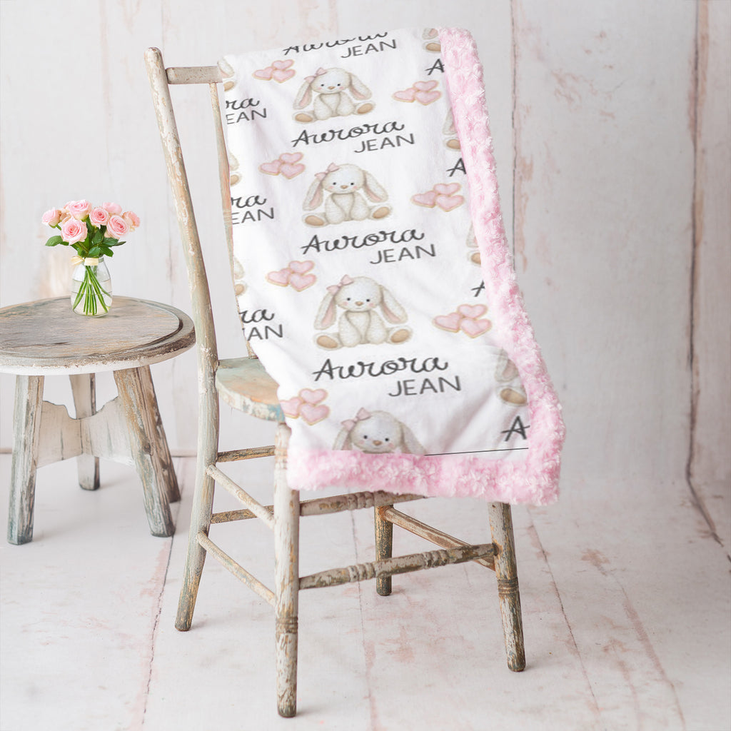 A personalized baby blanket draped over a rustic wooden chair, featuring a white background with a pattern of beige bunny illustrations, pink bows, and the text 'Aurora JEAN'. The blanket has a pink faux-fur trim.