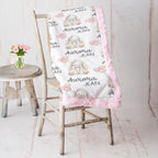 A personalized baby blanket draped over a rustic wooden chair, featuring a white background with a pattern of beige bunny illustrations, pink bows, and the text 'Aurora JEAN'. The blanket has a pink faux-fur trim.