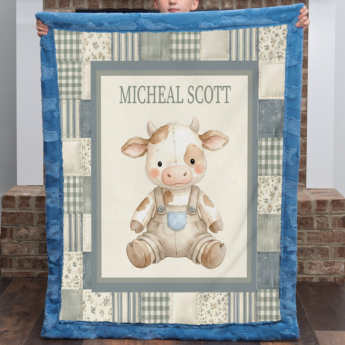 A personalized patchwork quilt held up by a child, featuring a central illustration of a cute cow in overalls with the name 'MICHEAL SCOTT' printed above it. The quilt has a blue border and a patchwork design with blue, beige, and gray tones.