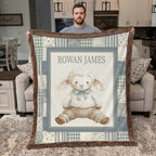 A man holds up a personalized patchwork-style baby blanket featuring a cartoon sheep illustration and the name 'ROWAN JAMES' in a real living room setting.