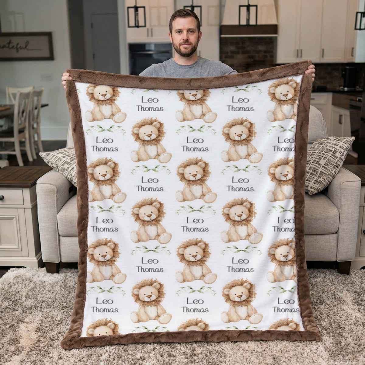 A man stands in a living room holding up a personalized blanket featuring a repeating pattern of teddy lions and the text 'Leo Thomas'.
