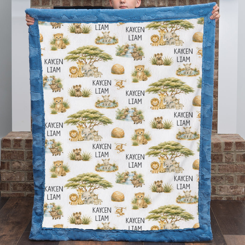A person holds up a minky quilt with a blue border and an all-over safari animal print featuring lions, giraffes, acacia trees, and the names 'KAYCEN LIAM'.