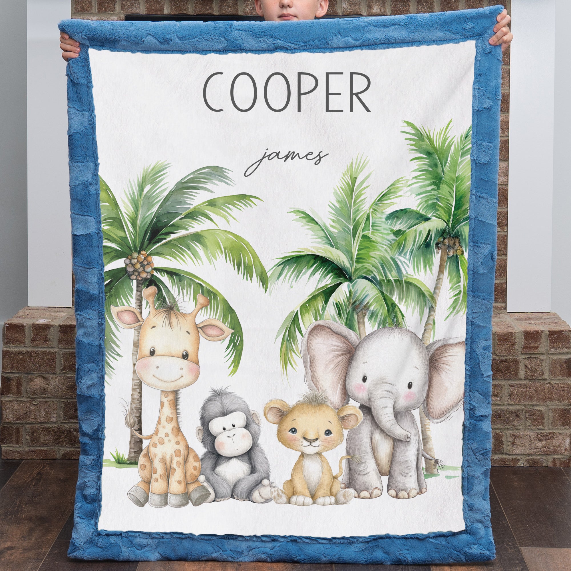 A personalized baby blanket with a blue ruffled border, featuring a white center printed with cartoon animals (giraffe, monkey, lion, elephant) and palm trees, with the names 'COOPER' and 'james' at the top. A person is holding it up in front of a brick wall.