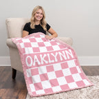 A woman sits in a beige armchair holding a pink and white checkered blanket with the name 'OAKLYNN' printed across the center. The scene is set in a room with a light gray wall, dark wood floor, and a shag rug.