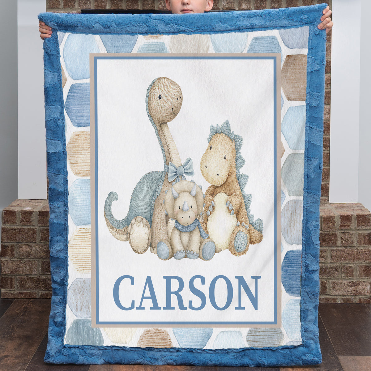 A personalized baby quilt held up by a child, featuring a blue ruffled border, a central illustration of three dinosaurs, and the name 'CARSON' printed at the bottom.