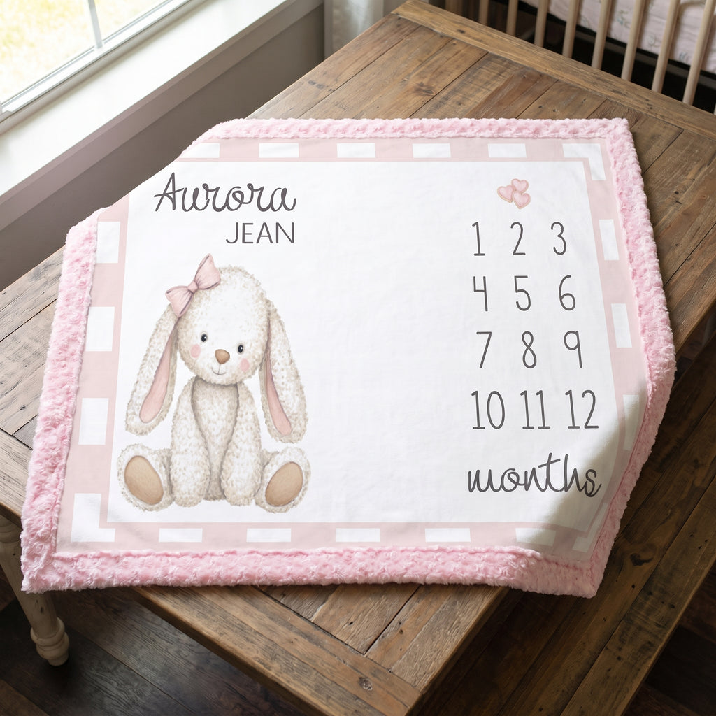 A personalized baby nursery blanket with a pink fuzzy border, featuring a white bunny illustration, the name 'Aurora Jean', numbers 1 through 12, and the word 'months' on a wooden table.