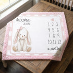 A personalized baby nursery blanket with a pink fuzzy border, featuring a white bunny illustration, the name 'Aurora Jean', numbers 1 through 12, and the word 'months' on a wooden table.