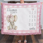 A person holds up a personalized baby blanket featuring a pink and white gingham border, a knitted elephant illustration with a pink bow, the name 'Eliana Elizabeth,' and a numbered month grid from 1 to 12. The blanket is shown in a home setting with a wood floor visible.