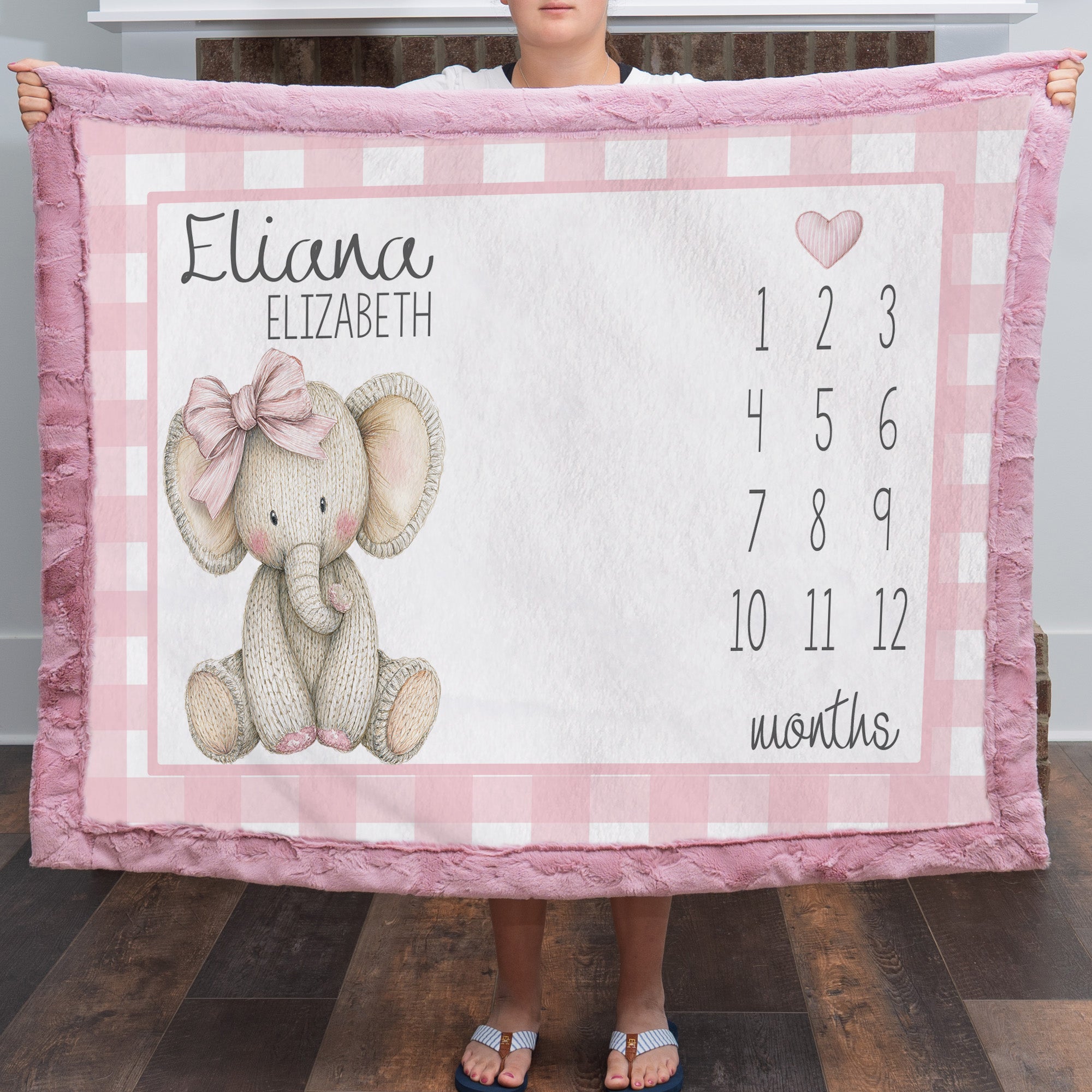 A person holds up a personalized baby blanket featuring a pink and white gingham border, a knitted elephant illustration with a pink bow, the name 'Eliana Elizabeth,' and a numbered month grid from 1 to 12. The blanket is shown in a home setting with a wood floor visible.