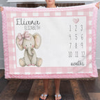 A person holds up a personalized pink and white checkered baby blanket featuring a knitted elephant illustration, the name 'Eliana Elizabeth,' and a months countdown grid.