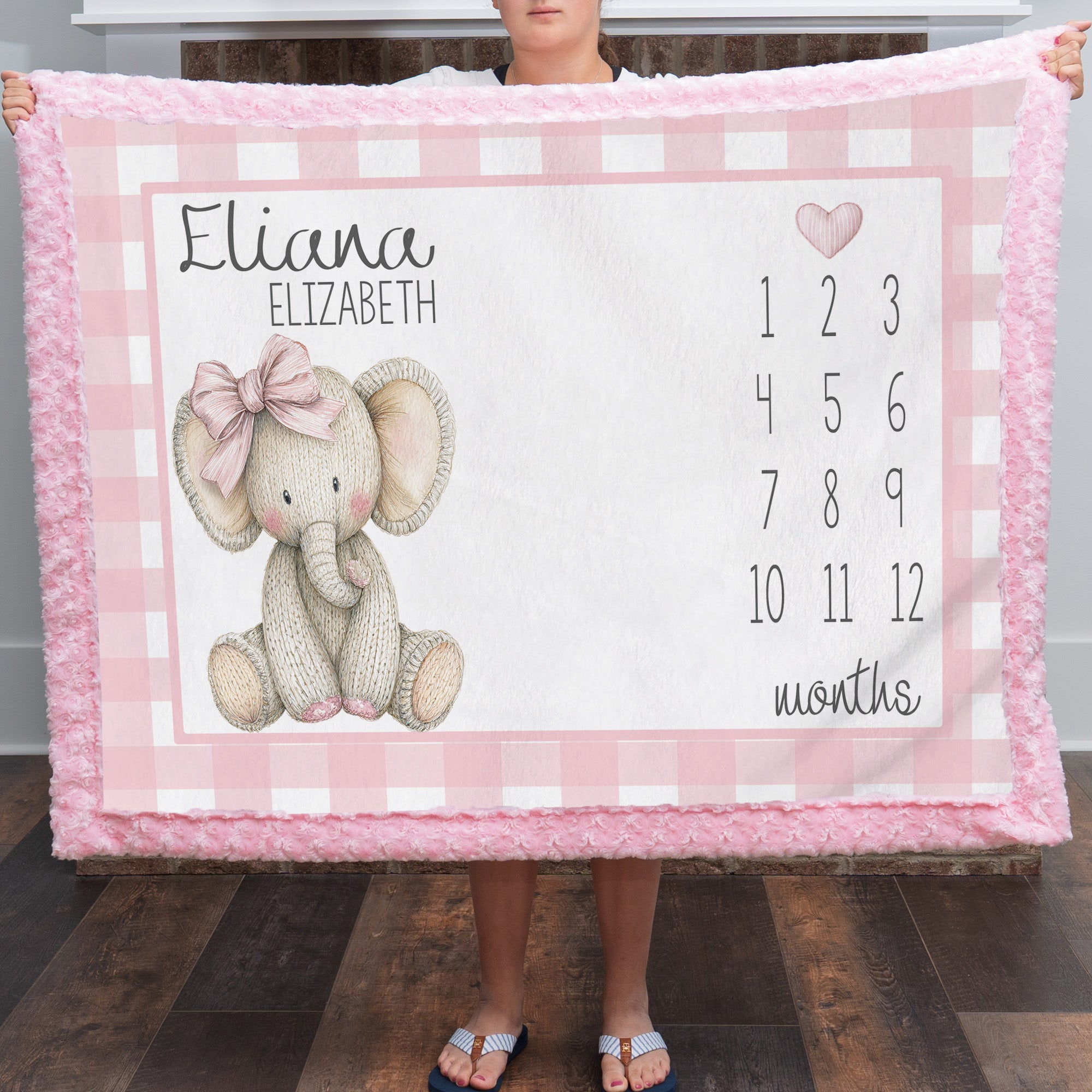 A person holds up a personalized pink and white checkered baby blanket featuring a knitted elephant illustration, the name 'Eliana Elizabeth,' and a months countdown grid.
