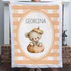 A person holds up a personalized quilt featuring a cute teddy bear sitting on a pumpkin, with the name 'GEORGINA' printed in the center. The quilt has a white border and a background pattern of peach and white stripes.