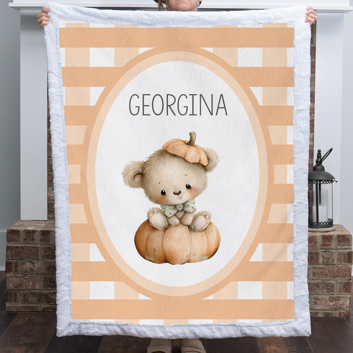 A person holds up a personalized quilt featuring a cute teddy bear sitting on a pumpkin, with the name 'GEORGINA' printed in the center. The quilt has a white border and a background pattern of peach and white stripes.