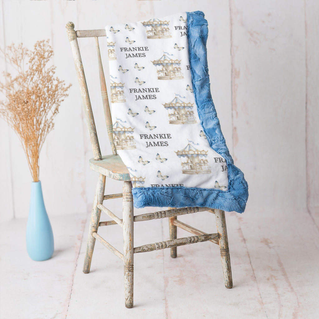 A personalized baby blanket with a white background, blue ruffled trim, and a carousel pattern with the name 'Frankie James', draped over a rustic wooden chair next to a light blue vase with dried grass.