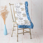 A personalized baby blanket with a white background, blue ruffled trim, and a carousel pattern with the name 'Frankie James', draped over a rustic wooden chair next to a light blue vase with dried grass.