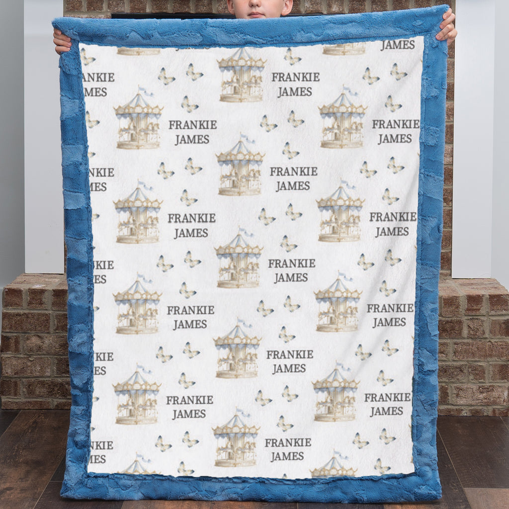A custom minky blanket held up by a child, featuring a white center printed with blue and beige carousel illustrations and the names 'Frankie James', surrounded by a blue plush border.